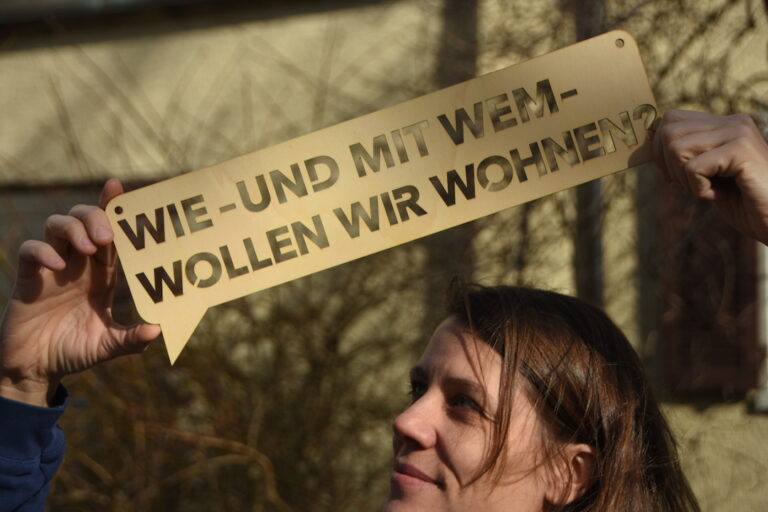 Frau mit Schild, darauf steht die Frage "Wie und mit wem wollen wir wohnen?"